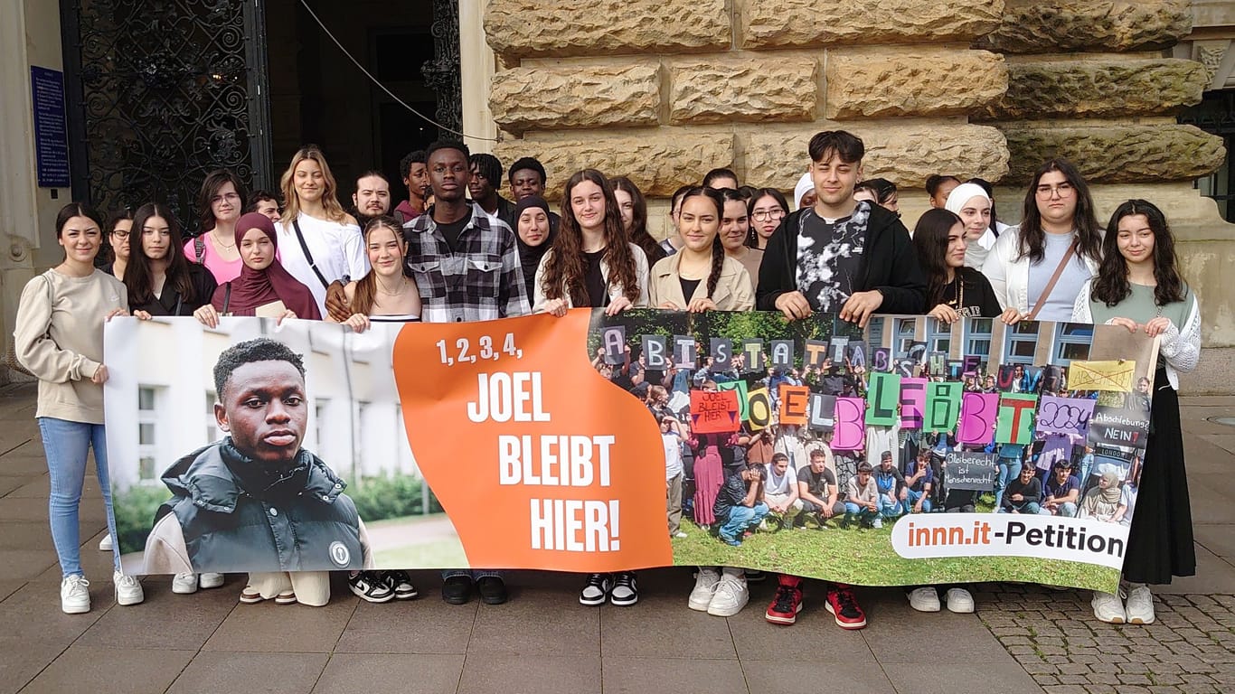 Joel aus Ghana (m.l., im grau-karierten Hemd) steht mit Mitschülern hinter einem Plakat mit der Forderung "1, 2, 3, 4 - Joel bleibt hier!" vor dem Rathaus.