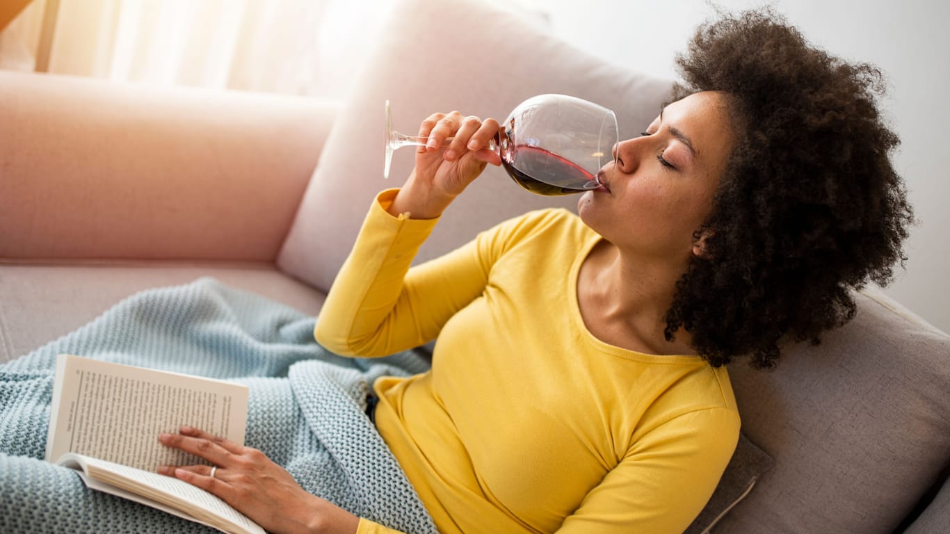 Frau sitzt entspannt mit einem Buch und einem Glas Wein auf dem Sofa