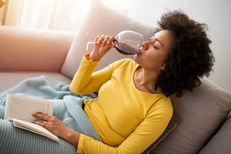Frau sitzt entspannt mit einem Buch und einem Glas Wein auf dem Sofa