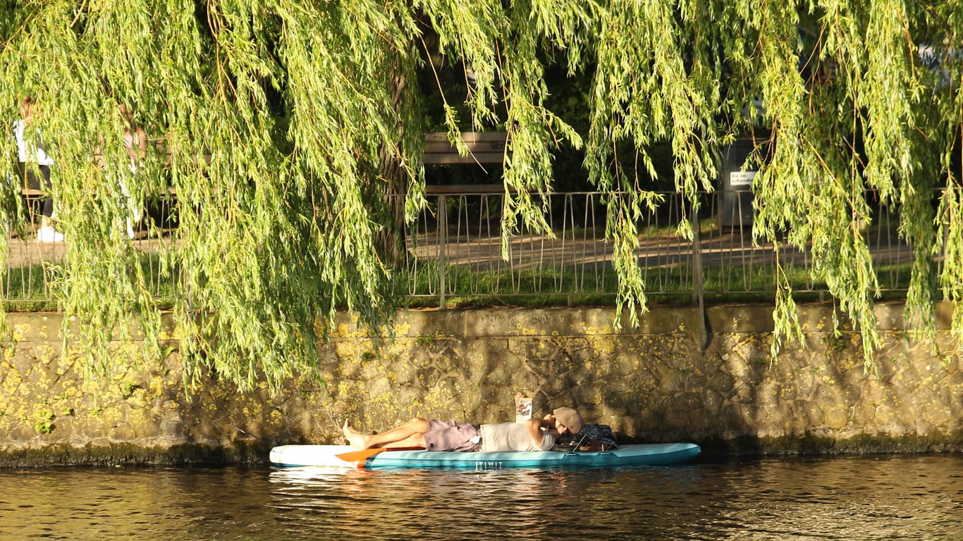 Ein Mann liegt auf einem Paddleboard auf einem Alsterkanal unter einem Baum: Am Sonntag sollte man sich besser nur im Schatten aufhalten.