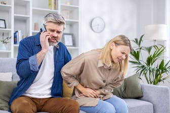 Ein Mann sitzt auf dem Sofa und telefoniert, neben ihm eine Frau mit Bauchschmerzen.