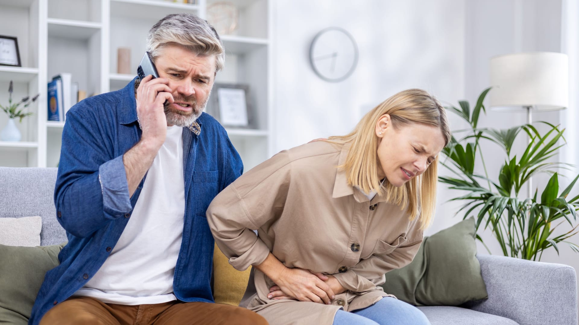 Ein Mann sitzt auf dem Sofa und telefoniert, neben ihm eine Frau mit Bauchschmerzen.