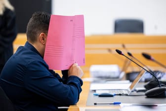 Der Angeklagte sitzt vor Verhandlungsbeginn in einem Gerichtssaal im Landgericht Hannover und hält sich eine Dokumentenmappe vor sein Gesicht. Neben ihm ist eine 41-Jährige angeklagt.