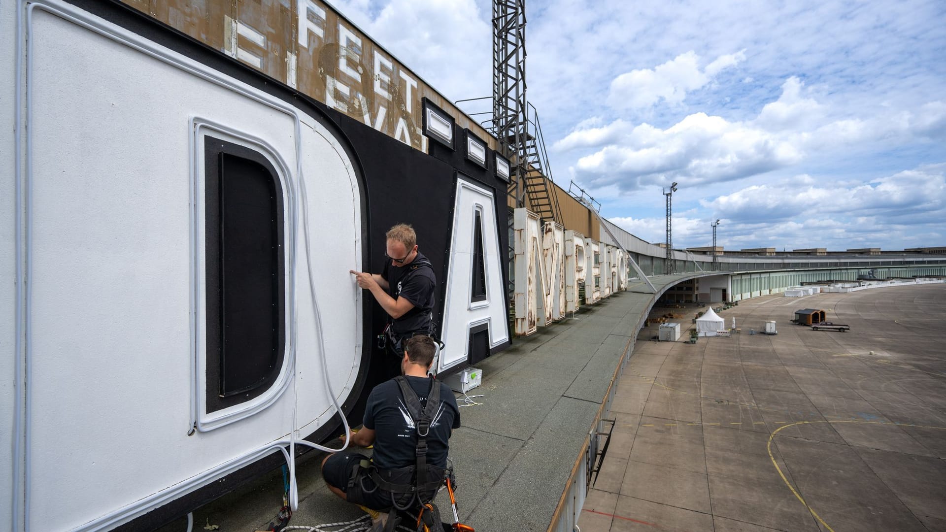 05.07.2024, Berlin: Zwei Mitarbeiter befestigen die Buchstaben des abgeänderten Schriftzuges "Berlin - DÄmpelhof" über dem ehemaligen Abfertigungsbereich des Flughafens Berlin-Tempelhof. Für einen Tag durfte die Band Die Ärzte im Vorfeld ihrer Konzerte am 23., 24. und 25. August 2024 den Schriftzug auf dem denkmalgeschützten Bau abändern