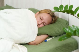Frau liegt krank im Bett, neben ihr ein Fieberthermometer
