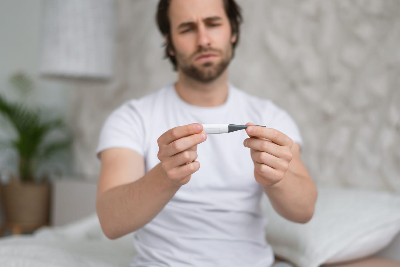 Mann sitzt auf dem Bett und schaut auf das Fieberthermometer