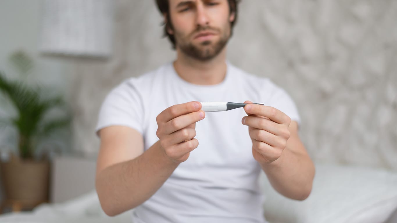 Mann sitzt auf dem Bett und schaut auf das Fieberthermometer