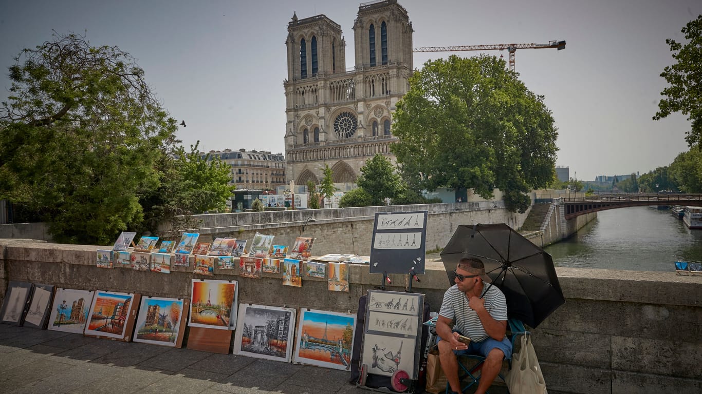 Ein Mann sitzt unweit der Notre-Dame mit einem Sonnenschirm (Archivbild): In den kommenden Tagen wird es in Paris heiß.