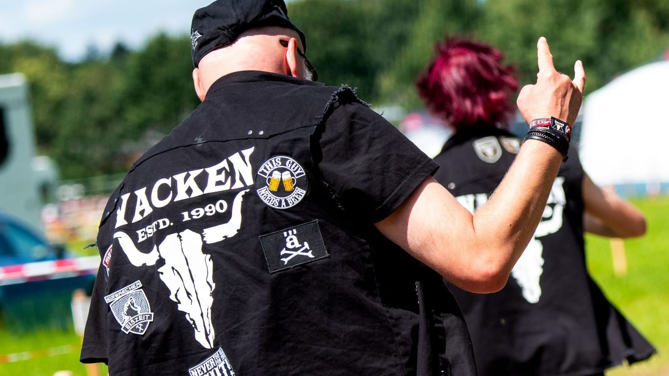 Ein Besucher vom Heavy-Metal-Festival Wacken Open Air geht über einen Campingplatz und zeigt mit gerecktem Zeigefinger und kleinem Finger den Wacken-Gruß.