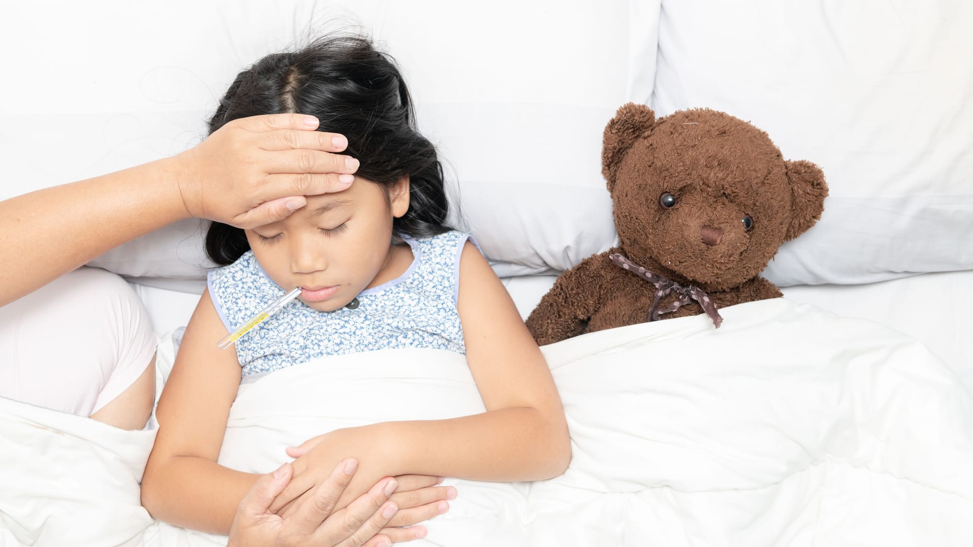 Kind liegt krank im Bett, die Mutter fühlt mit der Hand die Temperatur