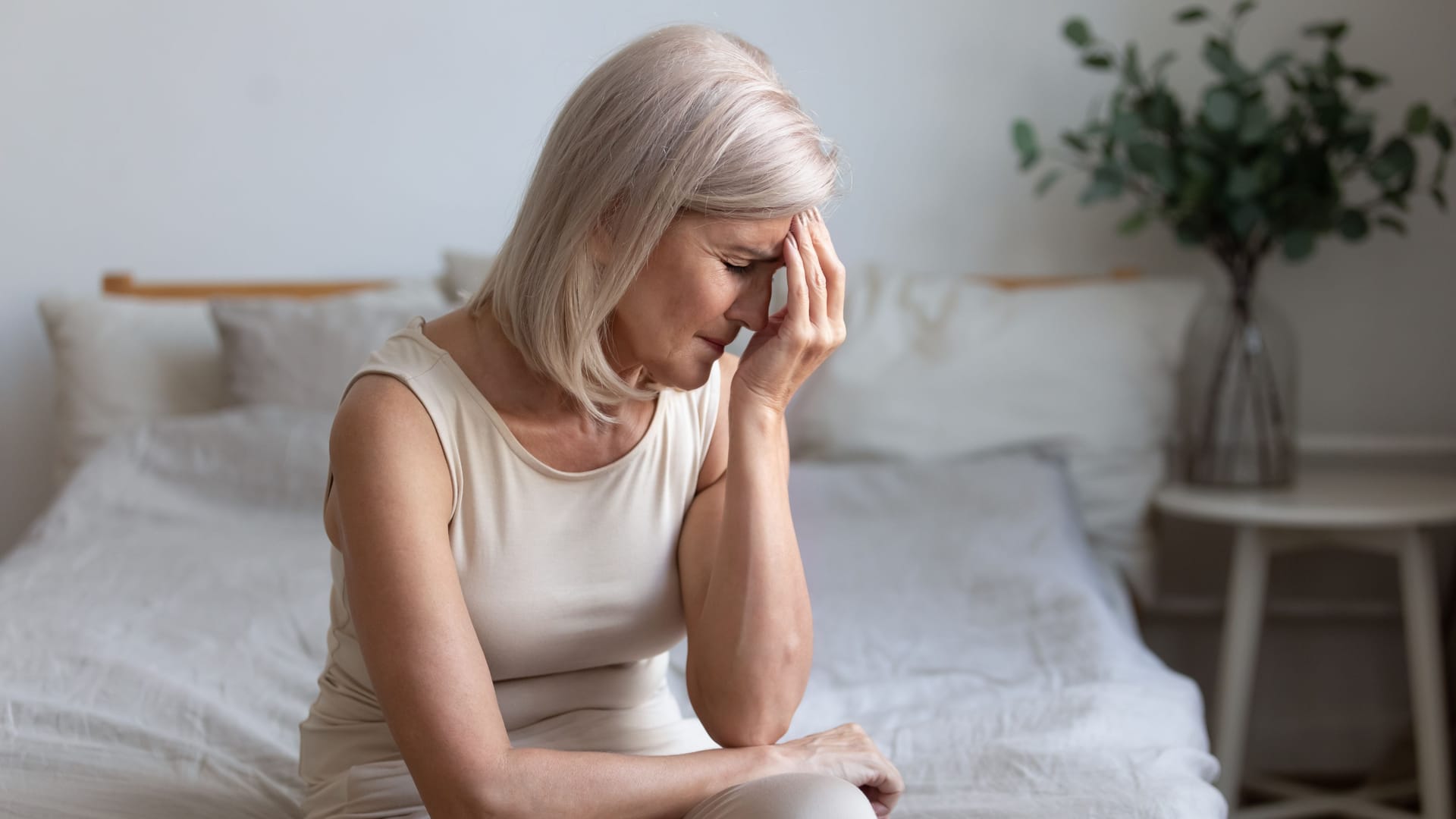 Frau sitzt auf dem Bett und fasst sich mit der Hand an die Stirn.