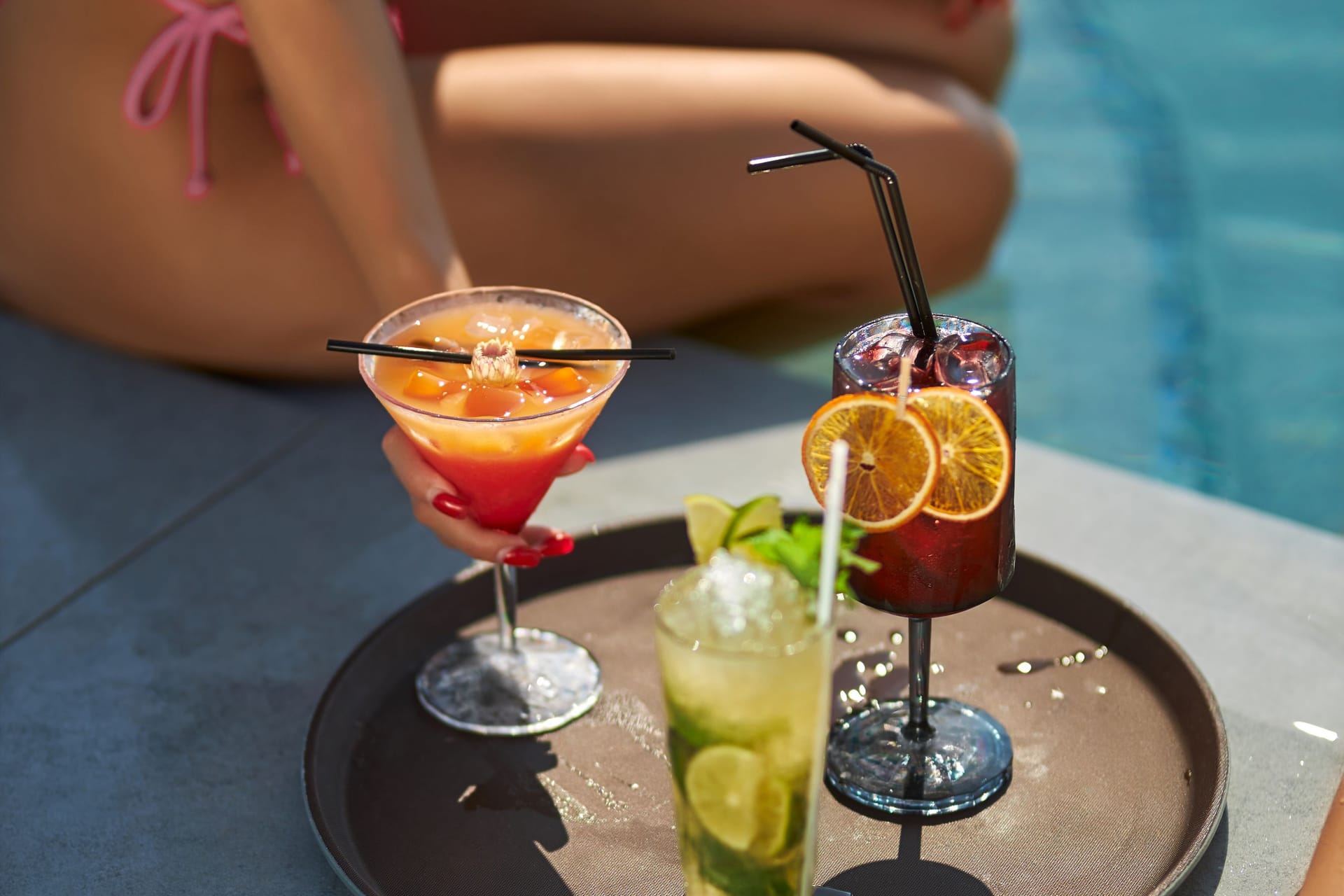 Tanned female in pink bikini drinking cocktails, while resting at poolside.