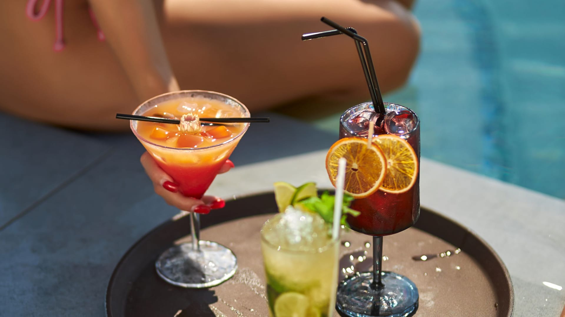 Tanned female in pink bikini drinking cocktails, while resting at poolside.