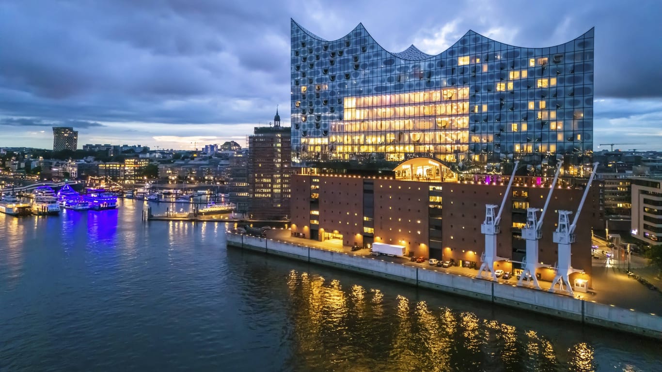 Das Bild zeigt die Elbphilharmonie in der Hamburger Hafencity am Abend zur sogenannten "blauen Stunde". Viele Lichter lassen das Gebäude erstrahlen. Der Himmel ist bewölkt und dunkel. Links ist ein blau leuchtendes Schiff zu sehen.