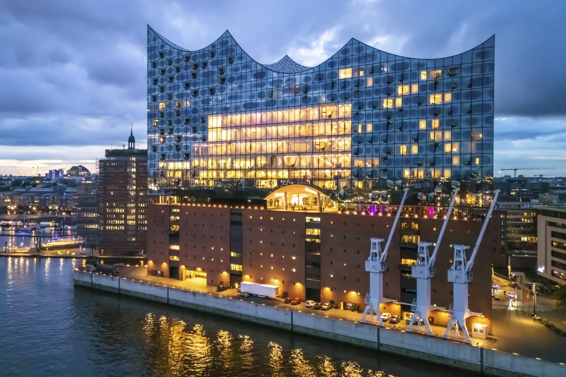 Das Bild zeigt die Elbphilharmonie in der Hamburger Hafencity am Abend zur sogenannten "blauen Stunde". Viele Lichter lassen das Gebäude erstrahlen. Der Himmel ist bewölkt und dunkel. Links ist ein blau leuchtendes Schiff zu sehen.