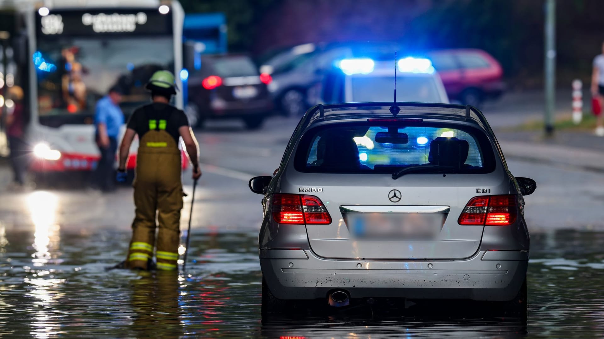 Nach dem Starkregen in Duisburg: Ein Auto steckt im Hochwasser in einer Senke fest, während Feuerwehrleute die Gullydeckel öffnen, damit das Wasser nach einem Unwetter ablaufen kann.