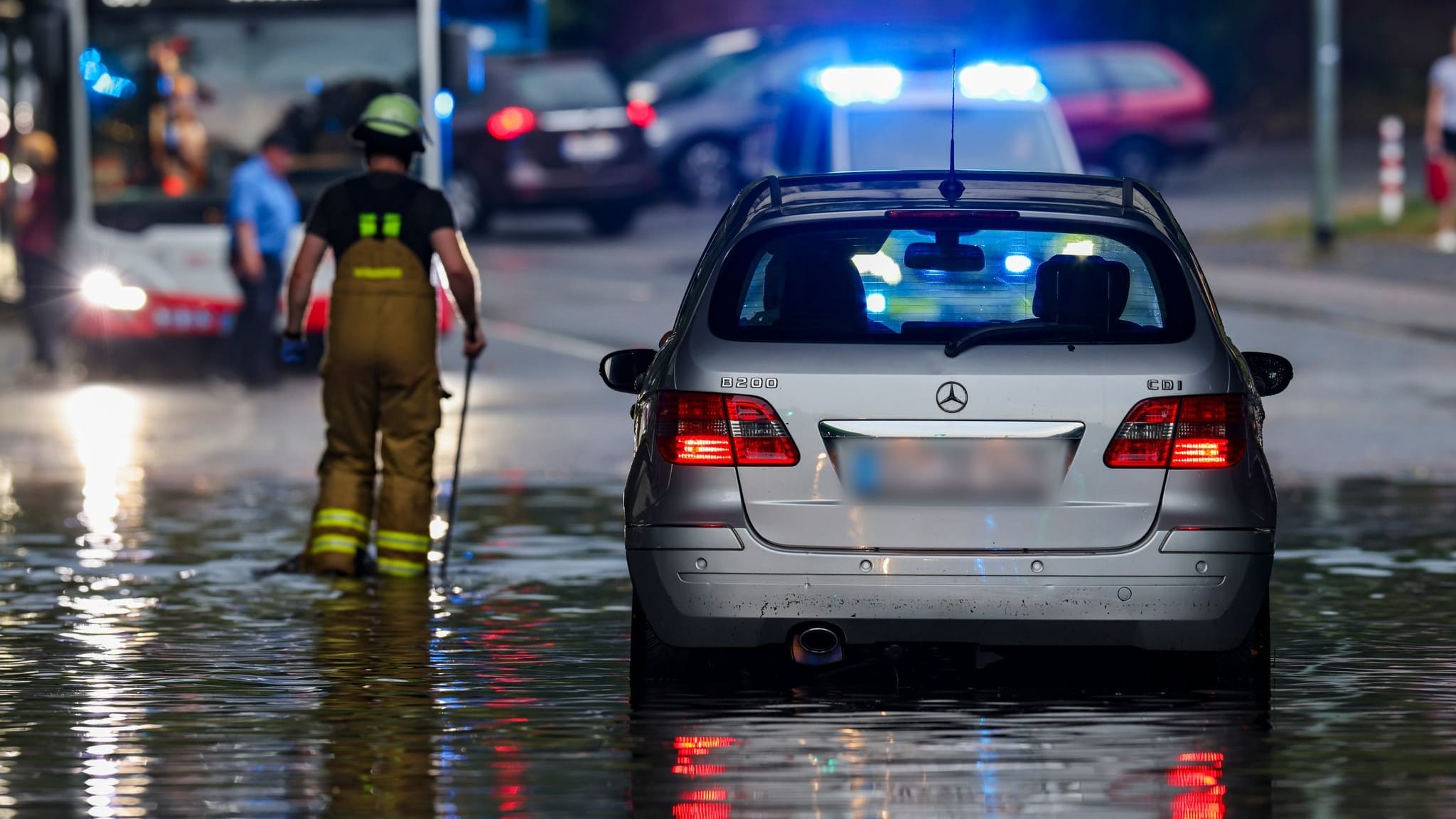 Nach dem Starkregen in Duisburg: Ein Auto steckt im Hochwasser in einer Senke fest, während Feuerwehrleute die Gullydeckel öffnen, damit das Wasser nach einem Unwetter ablaufen kann.