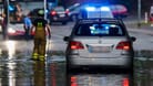 Nach dem Starkregen in Duisburg: Ein Auto steckt im Hochwasser in einer Senke fest, während Feuerwehrleute die Gullydeckel öffnen, damit das Wasser nach einem Unwetter ablaufen kann.