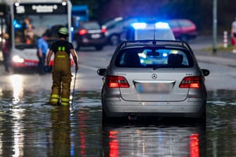 Nach dem Starkregen in Duisburg: Ein Auto steckt im Hochwasser in einer Senke fest, während Feuerwehrleute die Gullydeckel öffnen, damit das Wasser nach einem Unwetter ablaufen kann.