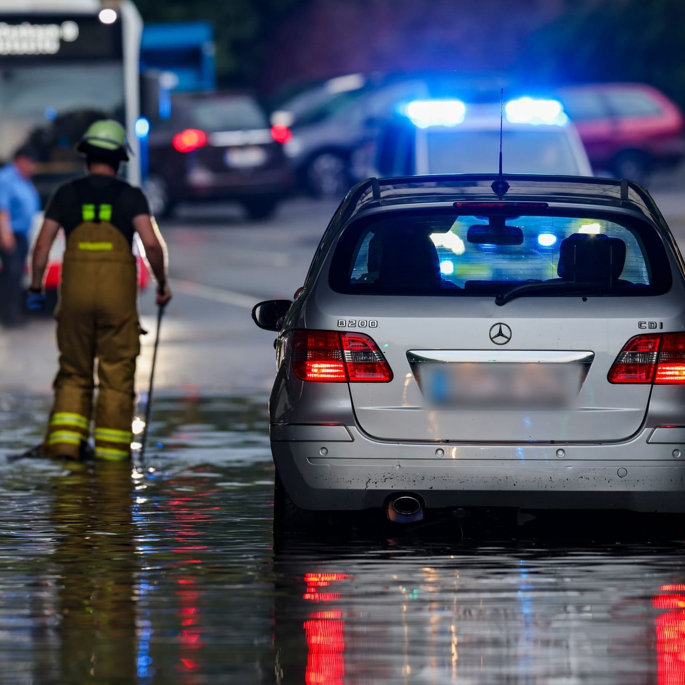 Nach dem Starkregen in Duisburg: Ein Auto steckt im Hochwasser in einer Senke fest, während Feuerwehrleute die Gullydeckel öffnen, damit das Wasser nach einem Unwetter ablaufen kann.