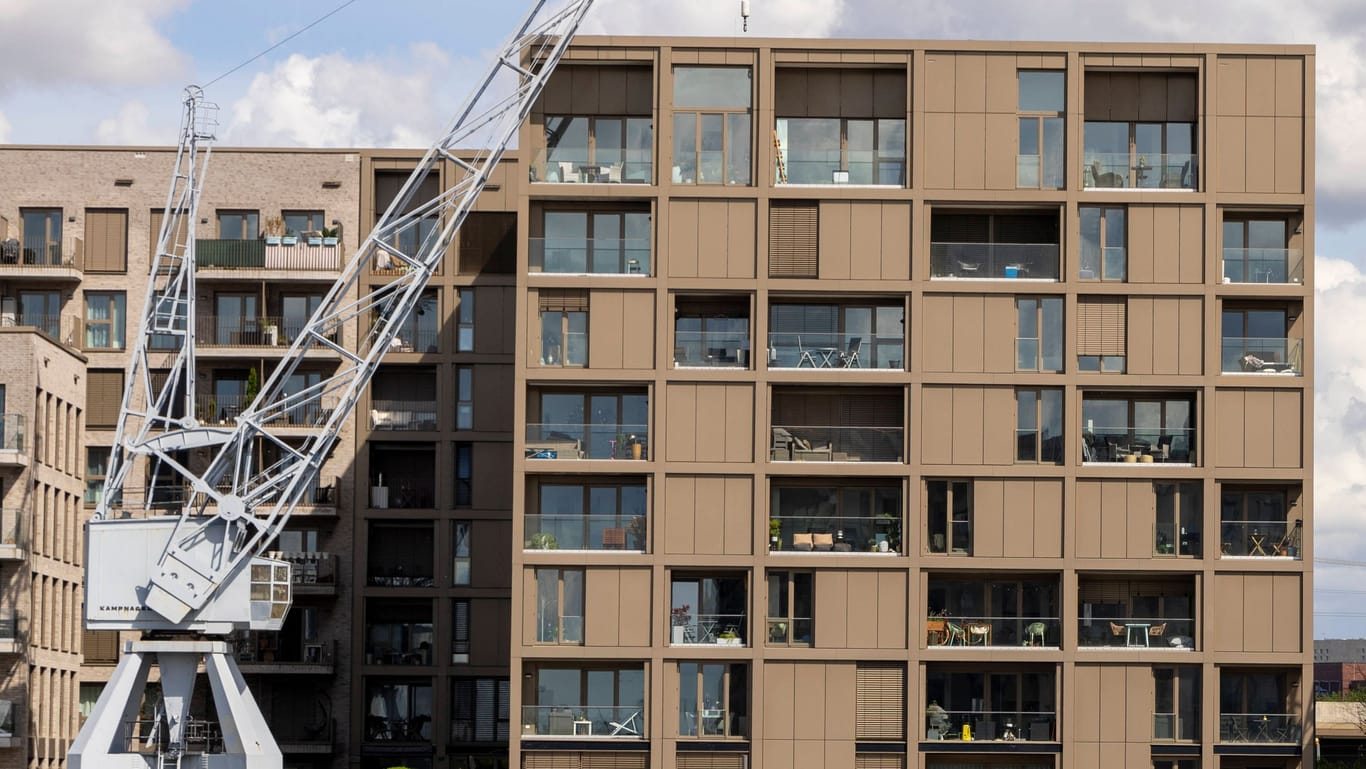 Das Bild zeigt neu gebaute Häuser am Baakenhafen in der Hafencity in Hamburg. Links stehen zwei braune Häuser, rechts ein weißes. Zu sehen ist jeweils eine große Fensterfront. Vor den Häusern stehen zwei weiße Kräne. Der Himmel ist bewölkt und blau.