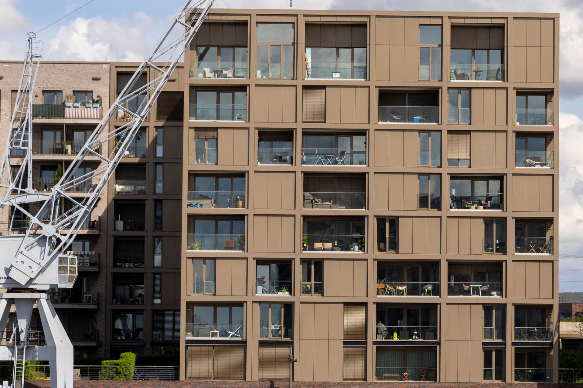 Das Bild zeigt neu gebaute Häuser am Baakenhafen in der Hafencity in Hamburg. Links stehen zwei braune Häuser, rechts ein weißes. Zu sehen ist jeweils eine große Fensterfront. Vor den Häusern stehen zwei weiße Kräne. Der Himmel ist bewölkt und blau.