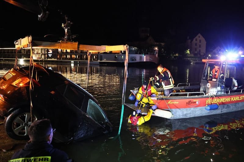 Die Feuerwehr birgt ein Auto in Karlstein am Main, das mit dem vorderen Teil über eine Kaimauer gefahren und im Wasser gelandet ist.