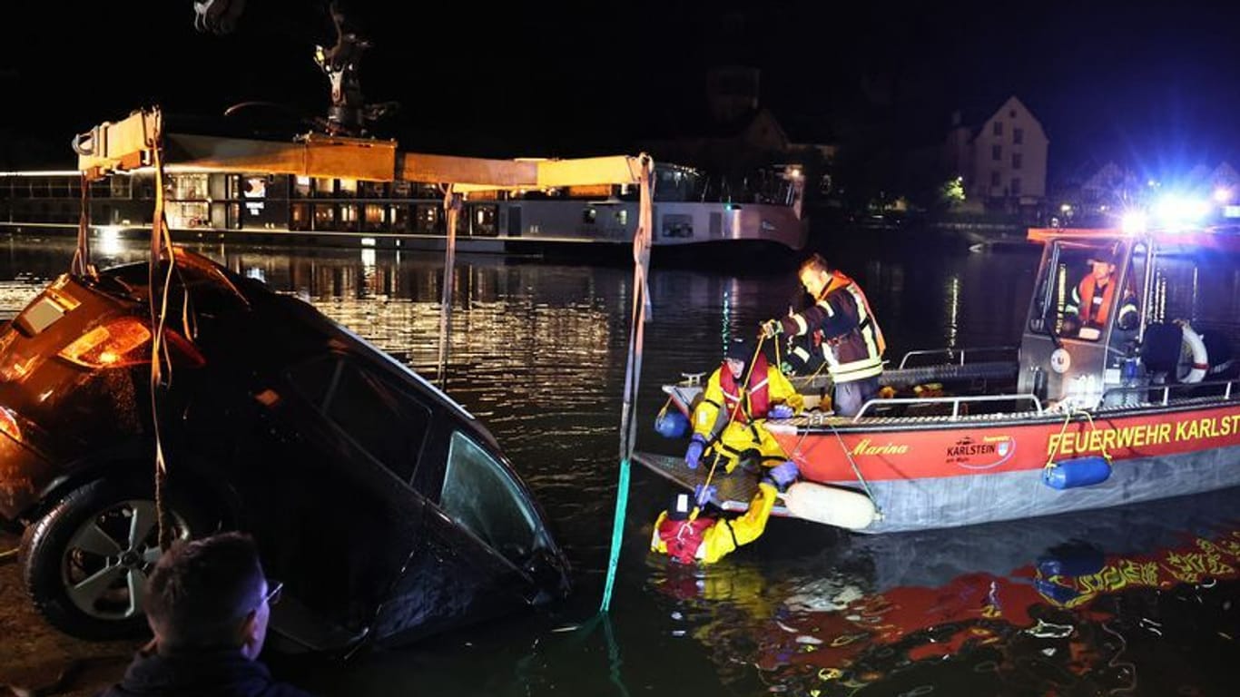 Die Feuerwehr birgt ein Auto in Karlstein am Main, das mit dem vorderen Teil über eine Kaimauer gefahren und im Wasser gelandet ist.