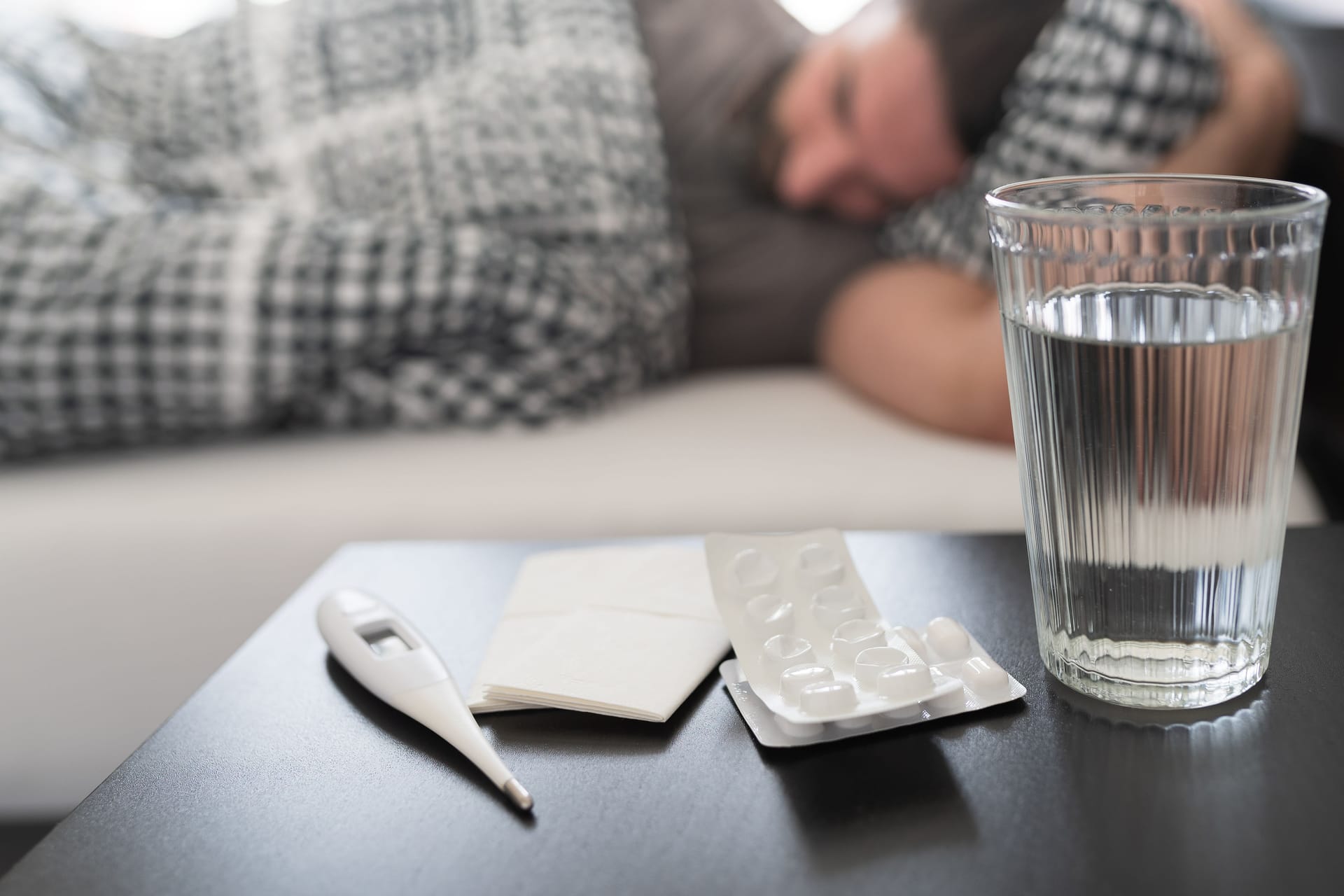 Mann liegt krank im Bett, im Vordergrund Tabletten, Wasserglas und Fieberthermometer