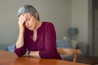 Frau sitzt am Tisch und hält sich eine Hand vor den Kopf.