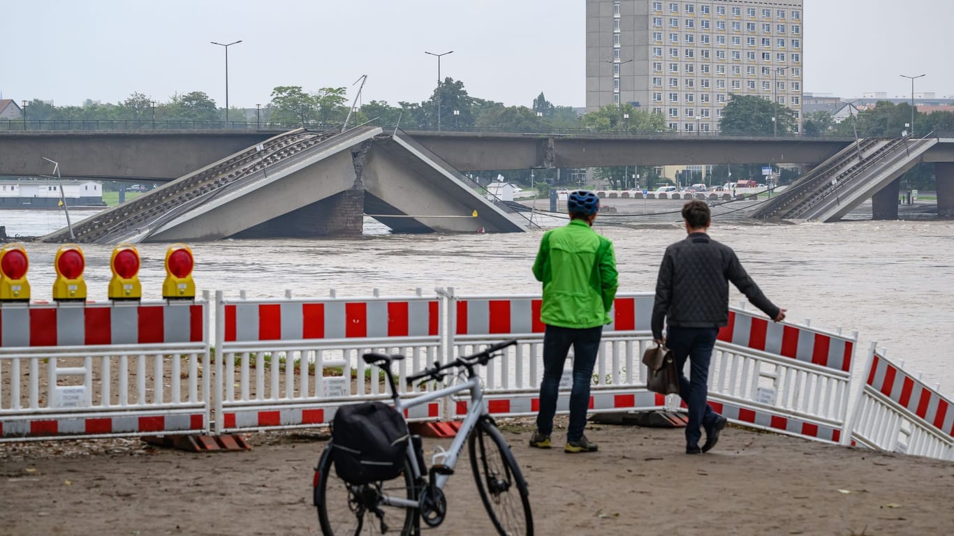 Zwei Männer stehen vor der vom Hochwasser der Elbe umspülten teileingestürzten Carolabrücke: Die Brücke ist weiträumig abgesperrt.