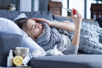 Kranke Frau liegt auf dem Sofa und schaut auf ein Fieberthermometer.