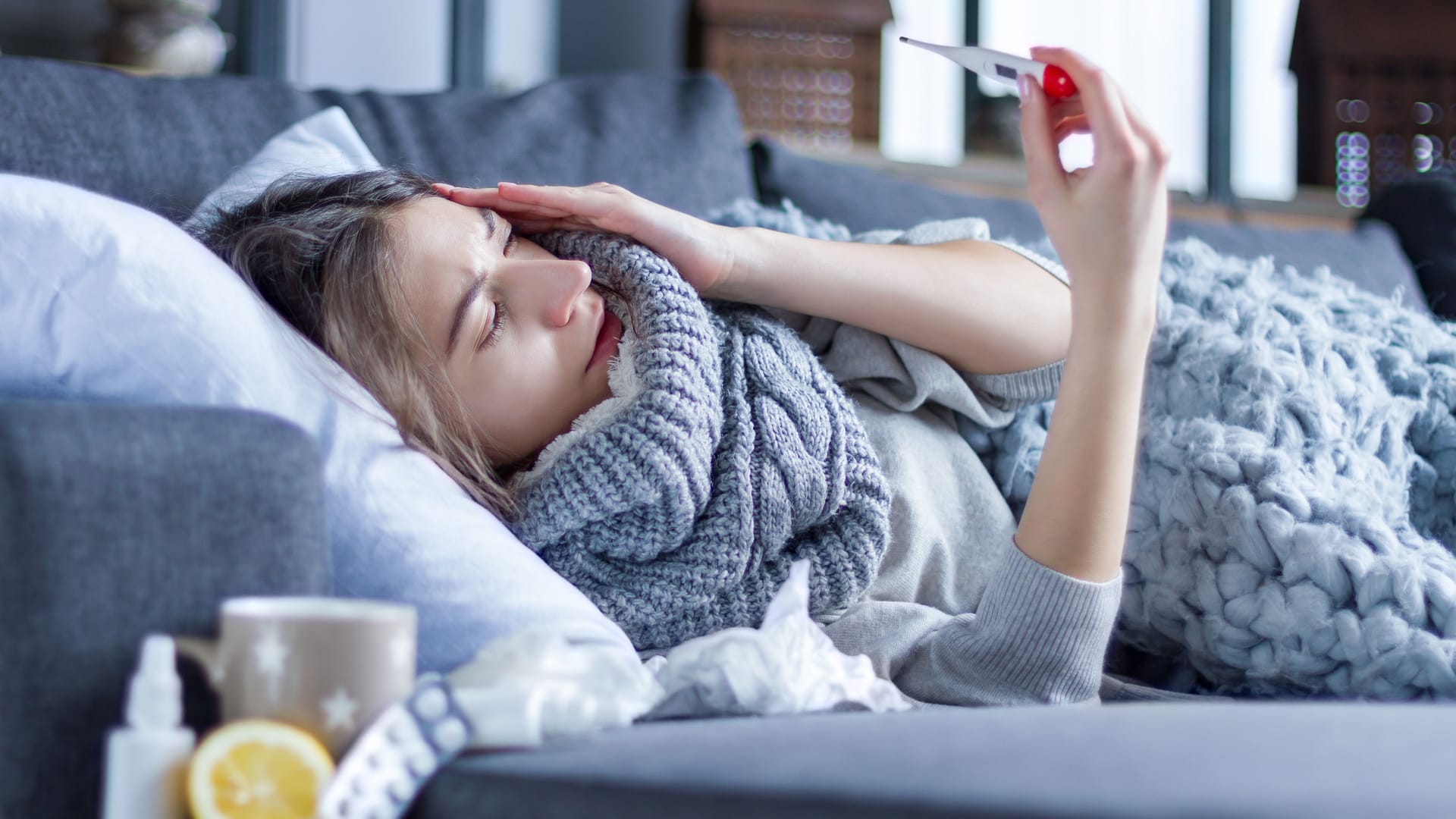 Kranke Frau liegt auf dem Sofa und schaut auf ein Fieberthermometer.