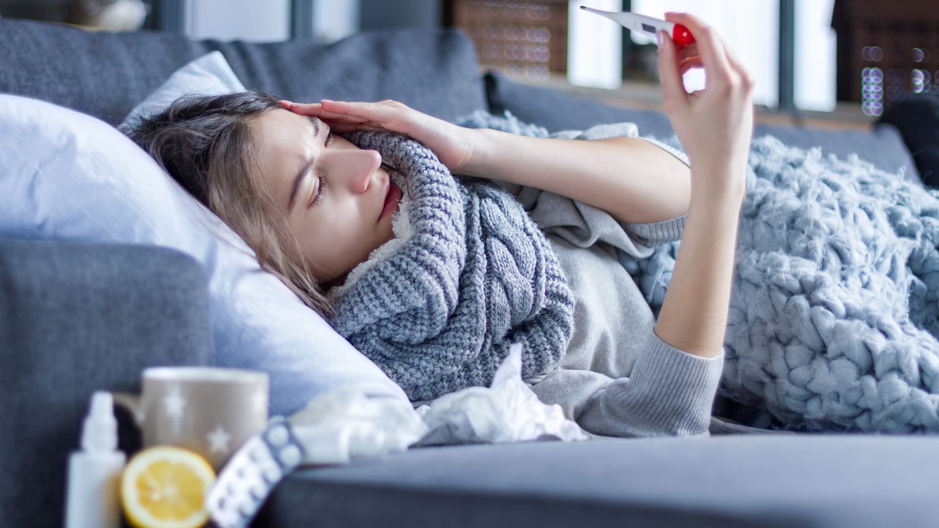 Kranke Frau liegt auf dem Sofa und schaut auf ein Fieberthermometer.