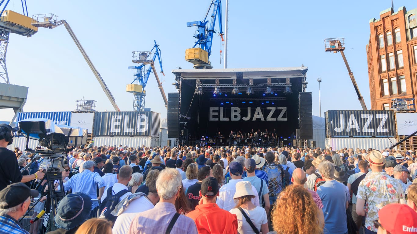 Zahlreiche Musikfans feiern am Hamburger Hafen auf dem Elbjazz-Festival (Archivbild). In der Bildmitte steht die Bühne, links im Hintergrund sind Kräne einer Werft zu sehen. Der Himmel ist blau, es scheint die Sonne.