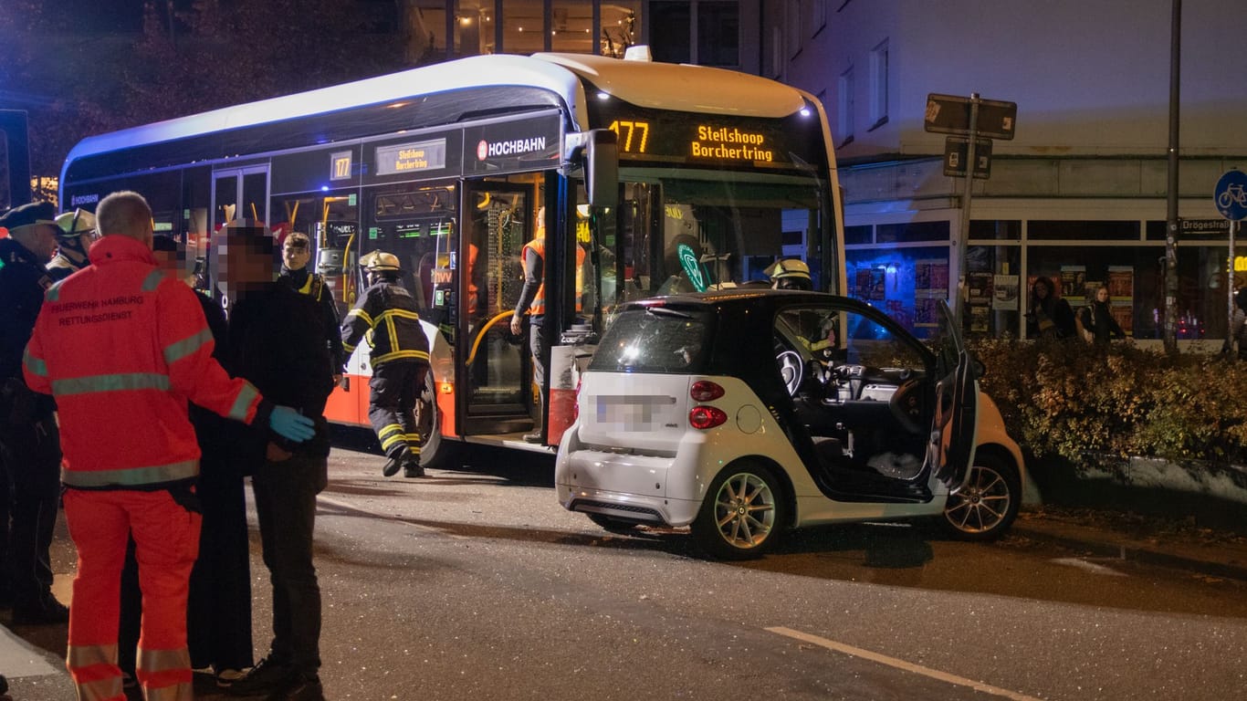 In Barmbek-Nord sind ein Auto und ein Bus kollidiert. Das kaputte, weiße Unfallauto steht auf der rechten Seite quer auf der Straße. Direkt dahinter steht der Linienbus. Feuerwehrleute laufen herum. Rettungskräfte kümmern sich um Unfallbeteiligte: