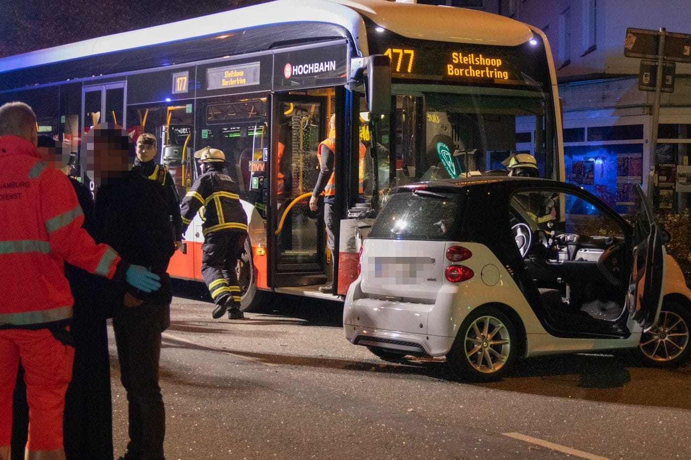 In Barmbek-Nord sind ein Auto und ein Bus kollidiert. Das kaputte, weiße Unfallauto steht auf der rechten Seite quer auf der Straße. Direkt dahinter steht der Linienbus. Feuerwehrleute laufen herum. Rettungskräfte kümmern sich um Unfallbeteiligte: