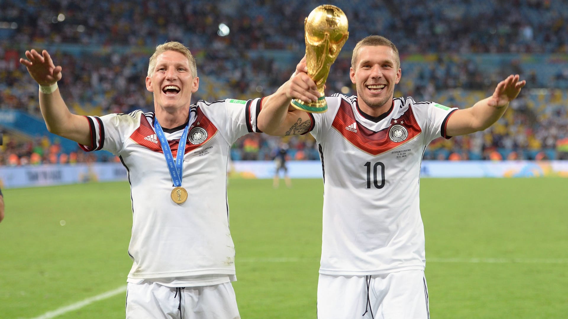 Lukas Podolski (r.) und Bastian Schweinsteiger bejubeln den Sieg der deutschen Nationalmannschaft im WM-Finale 2014. Sie machen mit ihren Händen eine Jubelpose und halten gemeinsam den WM-Pokal hoch. Sie tragen jeweils ein weißes Trikot der Nationalmannschaft. Im Hintergrund sind der Rasen des Stadions und Fans auf den Tribünen zu sehen.