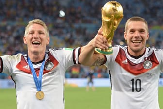 Lukas Podolski (r.) und Bastian Schweinsteiger bejubeln den Sieg der deutschen Nationalmannschaft im WM-Finale 2014. Sie machen mit ihren Händen eine Jubelpose und halten gemeinsam den WM-Pokal hoch. Sie tragen jeweils ein weißes Trikot der Nationalmannschaft. Im Hintergrund sind der Rasen des Stadions und Fans auf den Tribünen zu sehen.