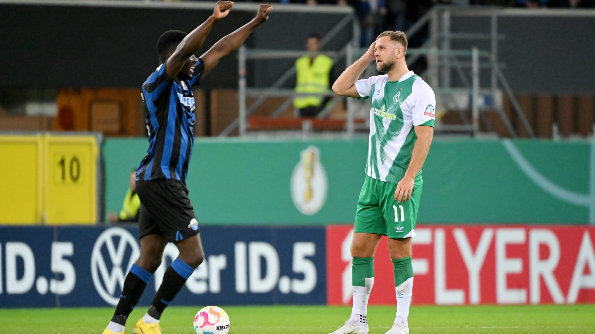 Paderborns Spieler Sirlord Conteh (l.) jubelt, er streckt die Arme gerade nach oben in die Luft. Rechts von ihm steht Werder Bremens Nicklas Füllkrug, der sich enttäuscht die rechte Hand an das Gesicht hält.