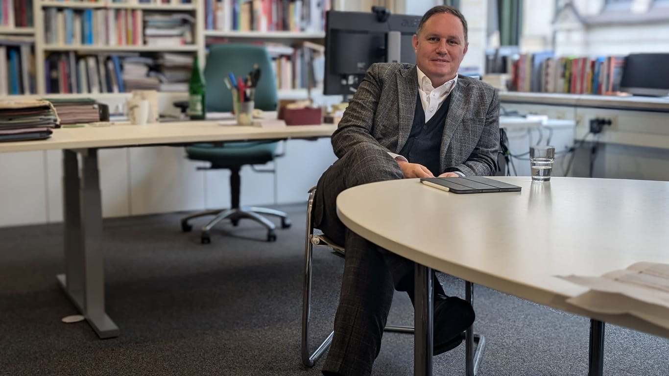 Hamburgs Kultursenator Carsten Brosda (SPD) sitzt in einem Büro an einem Schreibtisch in einem Stuhl. Er schlägt die Beine übereinander und schaut direkt in die Kamera. Auf dem Tisch liegt ein Tablet neben einem Wasserglas. Im Hintergrund sieht man Brosdas Arbeitsplatz und eine große Bücherwand.