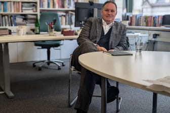 Hamburgs Kultursenator Carsten Brosda (SPD) sitzt in einem Büro an einem Schreibtisch in einem Stuhl. Er schlägt die Beine übereinander und schaut direkt in die Kamera. Auf dem Tisch liegt ein Tablet neben einem Wasserglas. Im Hintergrund sieht man Brosdas Arbeitsplatz und eine große Bücherwand.