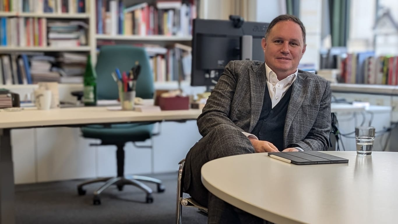 Hamburgs Kultursenator Carsten Brosda (SPD) sitzt in einem Büro an einem Schreibtisch in einem Stuhl. Er schlägt die Beine übereinander und schaut direkt in die Kamera. Auf dem Tisch liegt ein Tablet neben einem Wasserglas. Im Hintergrund sieht man Brosdas Arbeitsplatz und eine große Bücherwand.