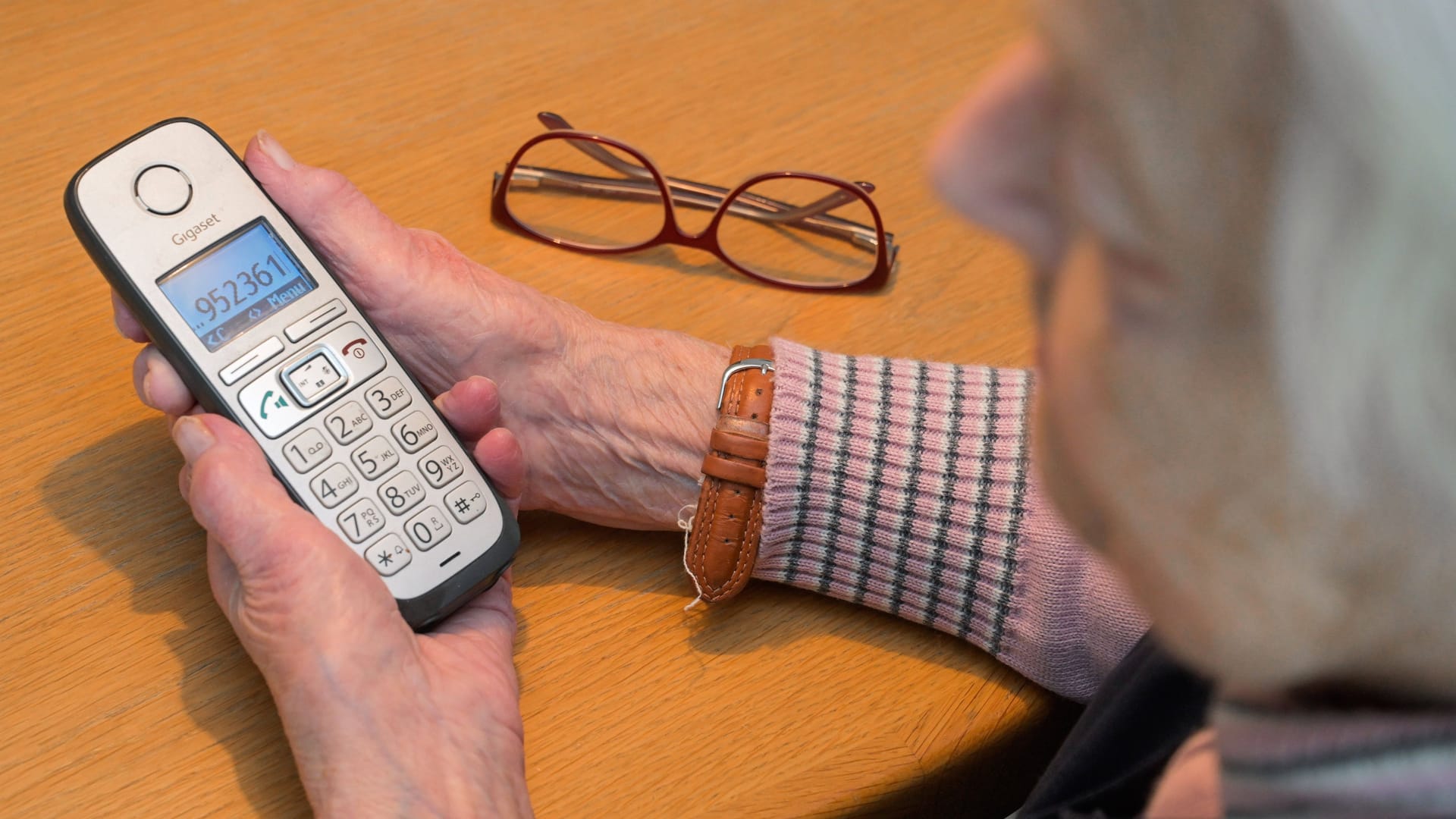 Eine ältere Frau hält ihr Festnetztelefon in den Händen. Auf dem Bildschirm ist eine Nummer zu erkennen. Neben dem Telefon liegt eine Brille auf einem Tisch.