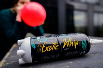 Eine Kartusche mit Lachgas der Sorte "Exotic Whip" liegt auf einem Tisch. Im Hintergrund zieht eine junge Frau aus einem roten Luftballon Lachgas.