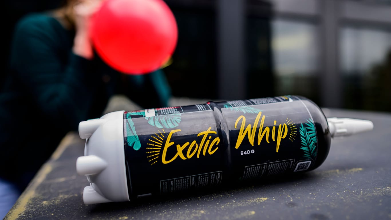 Eine Kartusche mit Lachgas der Sorte "Exotic Whip" liegt auf einem Tisch. Im Hintergrund zieht eine junge Frau aus einem roten Luftballon Lachgas.