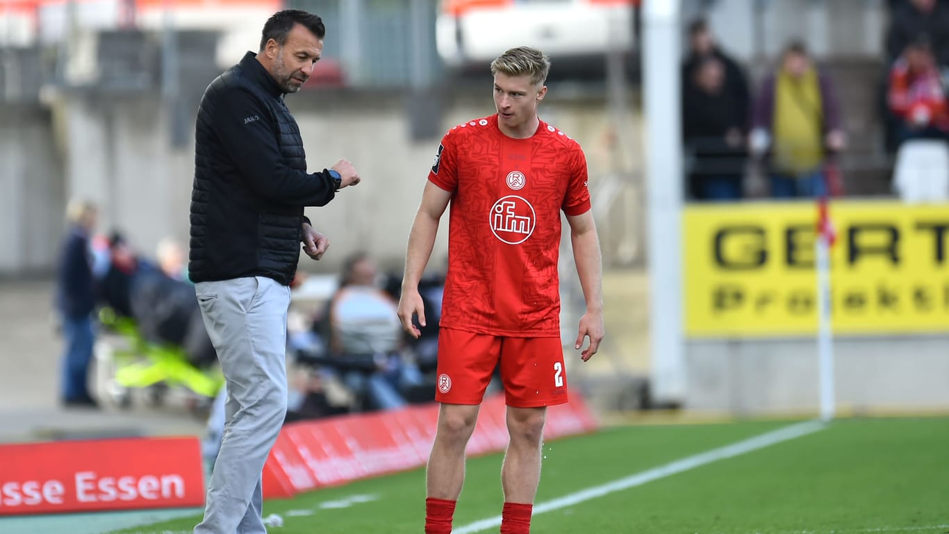 Rot-Weiss-Essen-Trainer Christoph Dabrowski (l) spricht mit seinem Spieler Julian Eitschberger während eines Spiels. Im Hintergrund sind Fans und Werbetafeln zu erkennen. Eitschberger trägt ein rotes Trikot, eine rote Hose und rote Socken.