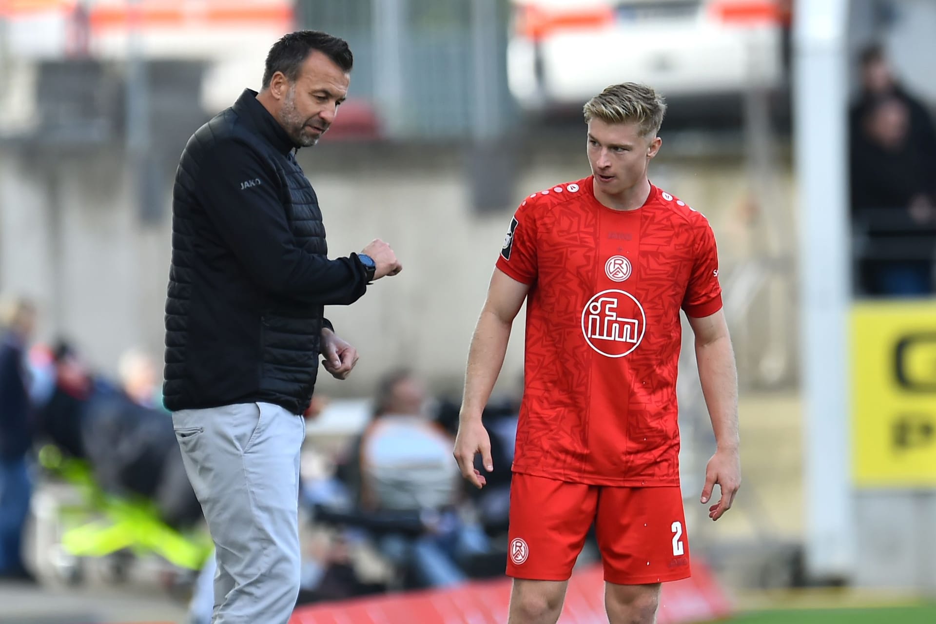 Rot-Weiss-Essen-Trainer Christoph Dabrowski (l) spricht mit seinem Spieler Julian Eitschberger während eines Spiels. Im Hintergrund sind Fans und Werbetafeln zu erkennen. Eitschberger trägt ein rotes Trikot, eine rote Hose und rote Socken.