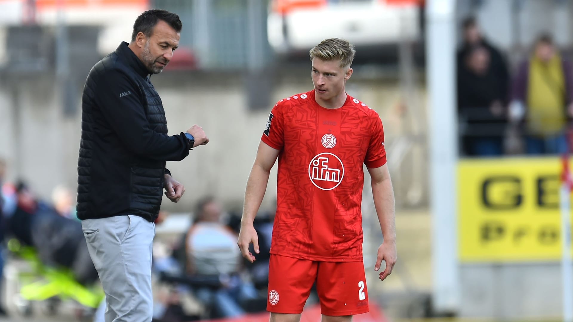 Rot-Weiss-Essen-Trainer Christoph Dabrowski (l) spricht mit seinem Spieler Julian Eitschberger während eines Spiels. Im Hintergrund sind Fans und Werbetafeln zu erkennen. Eitschberger trägt ein rotes Trikot, eine rote Hose und rote Socken.