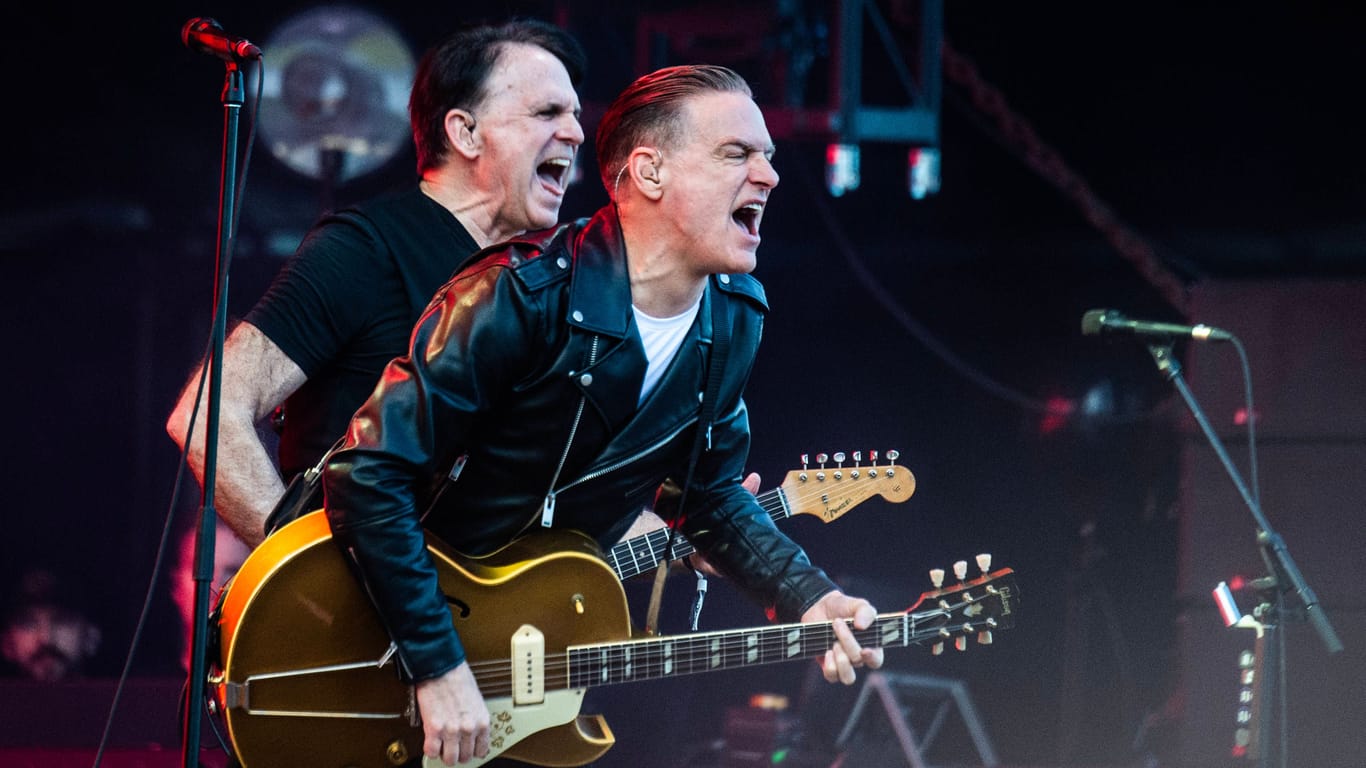 Bryan Adams (r.) und Keith Scott stehen auf einer Bühne. Beide Musiker spielen auf einer Gitarre und singen. Bryan Adams trägt eine schwarze Lederjacke, Keith Scott trägt ein schwarzes T-Shirt. Links und rechts von ihnen steht jeweils ein Mikrofon. Im Hintergrund sind Scheinwerfer zu erkennen.